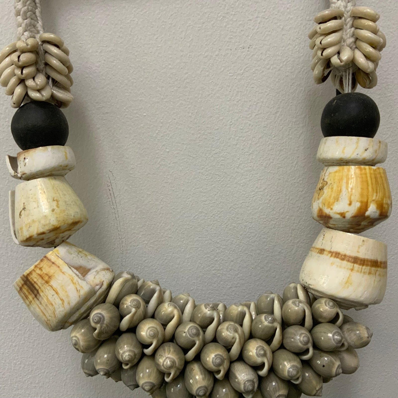 A handcrafted tribal-style statement necklace made of white sea shells, polished bone-colored resin rings, and black beads, displayed on a minimalist black metal stand.